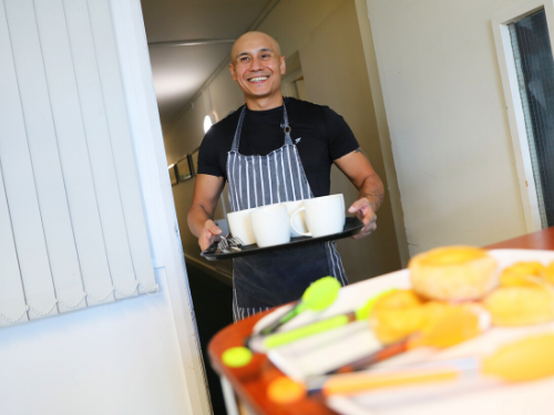 Smiling man with shaved head, black t-shirt and apron carrying tray of mugs in st vincent's newcastle